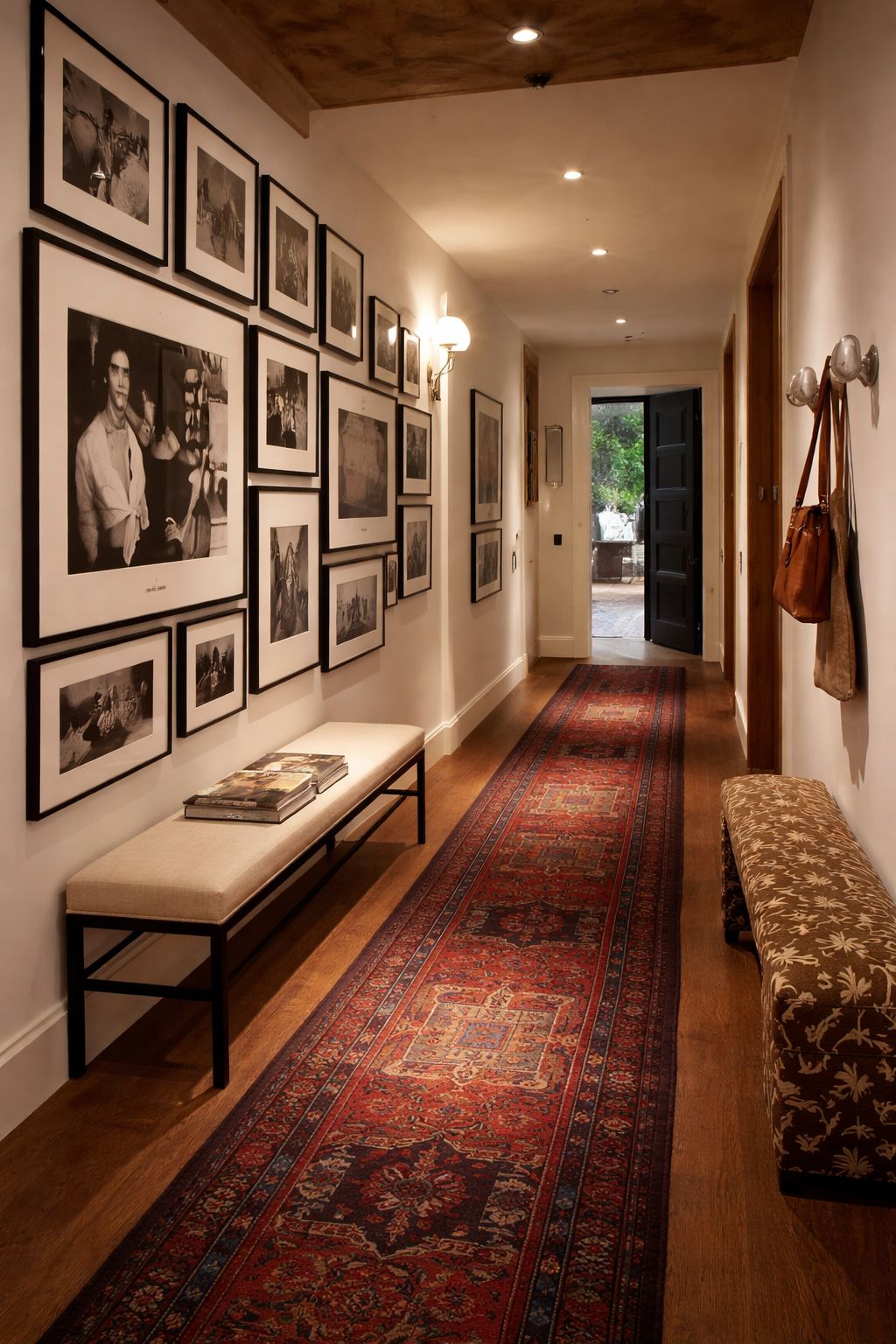 A hallway lined with a salon-style gallery wall of black-and-white framed photographs above an upholstered bench, with a Persian runner over warm timber floors.